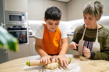Anne, küçük çocuğa mutfak tezgahında hamur yoğurmayı öğretiyor. Birlikte yemek hazırlıyorlar, aile bağlarını simgeliyorlar.