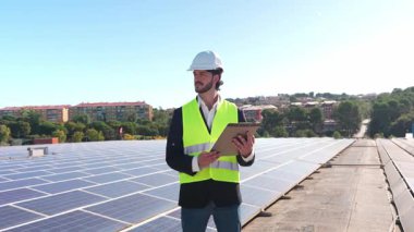 Sıkı bir şapka ve güvenlik yeleği giyen profesyonel bir mühendis elinde bir pano tutuyor ve çatıdaki bir güneş paneli kurulumunu inceliyor.