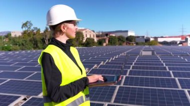 Sert bir şapka ve güvenlik yeleği giymiş kadın elektrik mühendisi dizüstü bilgisayarında çalışıyor, bir elektrik santralindeki güneş panellerini kontrol ediyor.