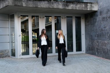 İki genç iş kadını iş ortaklığını ve başarıyı sembolize eden profesyonel bir ortamdan dışarı çıkıp konuşuyorlar.