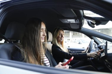 İki genç kadın modern bir araç kullanıyor, gösterge panelinde navigasyon kullanıyor ve elinde cep telefonu olan bir yolcu.
