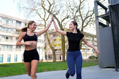 İki gülümseyen bayan arkadaş parkta koşu yaparken beşlik çakıyor takım çalışmasını ve fitness hedeflerini kutluyorlar.