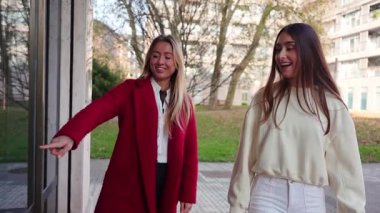 Birlikte alışveriş yapan iki şık kadın. Kırmızı ceketli sarışın kadın çantadan arkadaşına sarı bir tişört gösteriyor.