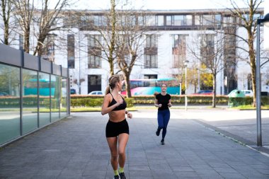 Şehir parkında kaldırımda koşmaktan keyif alan iki genç kadın sağlık ve fitness yaşam tarzını destekliyor.