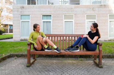Spor yapan iki arkadaş bir bankta dinleniyor, spor yaptıktan sonra dışarıda canlı sohbetler ediyorlar.