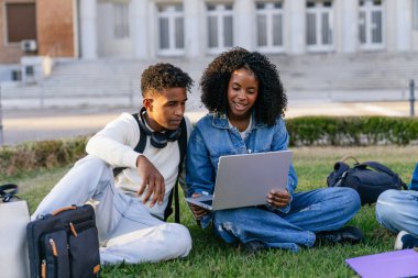 Afrika kökenli Amerikalı çift bir dizüstü bilgisayarla çalışıyor, bir üniversite binasının önünde çimlerin üzerinde oturuyorlar.