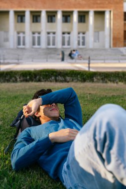 Genç adam yeşil çimlerde dinleniyor, gözleri güneşten koruyor, üniversite binası ve arka planda bulanık öğrenciler var.