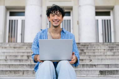 Üniversite merdivenlerinde elinde dizüstü bilgisayarla oturan mutlu öğrenci. Kampüste eğitim, teknoloji ve öğrenimi temsil etmek