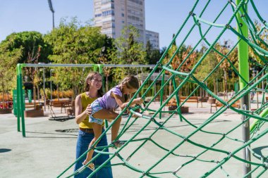Çocuk gelişimi ve şehir parkında aktif oyun için yeşil bir iple tırmanma ağında genç bir kıza yardım eden ebeveyn