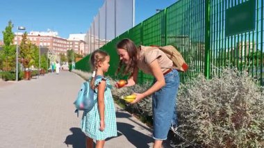 Genç anne kızına okula yürümeden önce sağlıklı bir meyve veriyor, sonra onu öpüyor ve güneşli bir günde el sallıyor.
