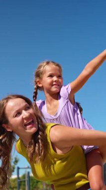 Neşeli küçük kız güneşli bir günde annesiyle eğleniyor. Genç bir kadın açık mavi gökyüzüne karşı sevimli çocuğunu sırtında taşıyor.