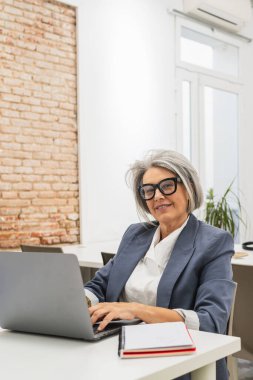 Kıdemli iş kadını kameraya gülümsüyor, parlak bir ofiste çalışırken dizüstü bilgisayarda yazıyor, kendine güvenini gösteriyor.