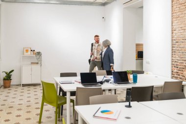 İki iş arkadaşı laptoplar ve masalarla dolu çağdaş ve aydınlık bir ofiste yürürken iş konuşuyorlar.