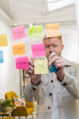 Girişimci adam, modern ofis alanındaki şeffaf bir duvara renkli yapışkan notlarla fikirler ve stratejiler üretiyor.