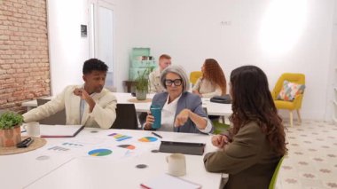 Modern bir ofiste yapılan bir toplantıda, çok nesilli ve çok ırklı işadamları çizelge ve verileri kağıt üzerinde analiz edip tablet hazırlıyorlar.