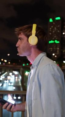 Sarı kulaklıklı neşeli adam akıllı telefonuyla müzik dinliyor. Şehir köprüsünde gece hayatı ile dans eden neşeli adam.