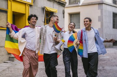 Gururu kutlayan bir grup genç arkadaş, ellerinde gökkuşağı bayrakları ve aksesuarlarıyla bir şehir caddesinde, mutluluk ve kapsamı ifade ediyorlar.