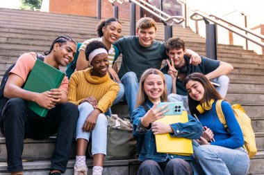 Çeşitli gülümseyen öğrenciler üniversite merdivenlerinde oturup selfie çekiyorlar, keyifli bir anı paylaşıyorlar.
