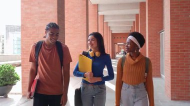 Üç mutlu çok kültürlü üniversite öğrencisi tuğla sütunlarda birlikte yürüyorlar. Sınıfa giderken konuşan ve gülümseyen genç arkadaşlar.