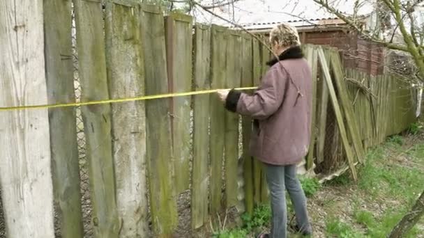 Деревенская девушка измеряет старый забор рулеткой — стоковое видео