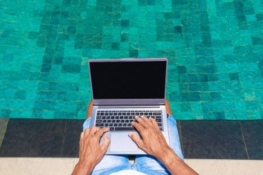 Bir adam havuz başında dizüstü bilgisayarda çalışıyor. Serbest çalışmak, doğada çalışmak. Eller yazı yazıyor..