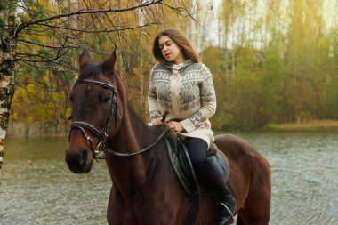 Güz ormanında göl kenarında at sırtında tatlı bir genç kadın. Sürücü dişi, yağmurlu bulutlu bir havada atını Park 'ta sürüyor. Açık hava biniciliği, spor ve eğlence anlayışı. Boşluğu kopyala