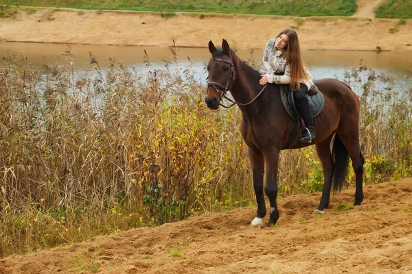Cute young woman on horseback in autumn forest by lake. Rider female drives her horse in Park in inclement cloudy weather with rain. Concept of outdoor riding, sports and recreation. Copy space
