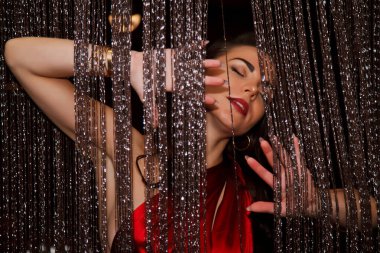 Kırmızı kokteyl elbisesi içinde oldukça seksi bir esmer gümüş iplik perdesinin arkasından dışarı bakıyor. Kadın flörtü ve aktrisin duyguları. Oyun ve flört kavramı. Boşluğu kopyala