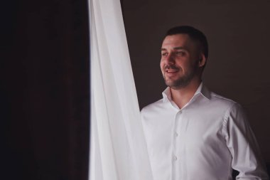 Yakışıklı genç damat, otel odasının penceresinde beyaz gömlek giyer. Gülümser gibi gülümser. Şirin çocuk gelinle tanışmaya hazırlanıyor. Mutluluk ve lüks evlilik kavramı. Boşluğu kopyala