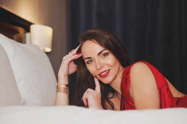 Otel yatağında yatan kırmızı elbiseli, genç ve seksi esmerin yakın çekimi. Flört eden aktrisin kadınsı duyguları. Sevgililer Günü için rahatlama, memnuniyet ve ilham. Boşluğu kopyala