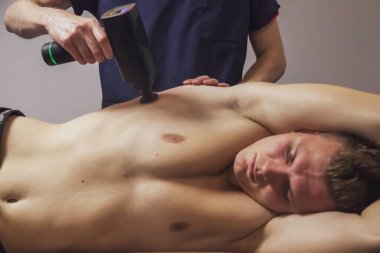 Masaj terapisti, profesyonel bir erkek sporcunun yaralanmasını tedavi ediyor. Spor salonunun tıbbi ofisinde spor tabancası şok masajı. Atletik vücut masajı için perküsyon terapisi. Fizyoterapi