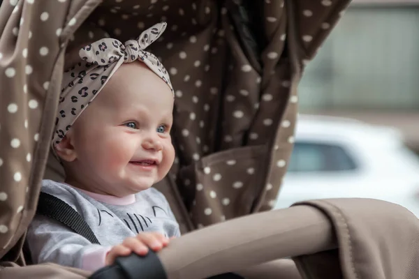 Happy smiling emotional eight-month-old blue-eyed girl sit in stroller on walk and wait for mom. Small and cute baby with headband on head sitting in pram. Concept of proper upbringing and childhood