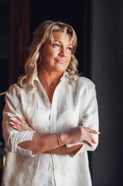 Orta yaşlı, sevimli, orta yaşlı, hizmetçi odasında sarışın bir kadın. Beyaz elbiseli, koyu renk arka planlı bir kadın. Pencerenin önünde yalınayak. Kapalı alanda kendinden emin olgun bir kadın