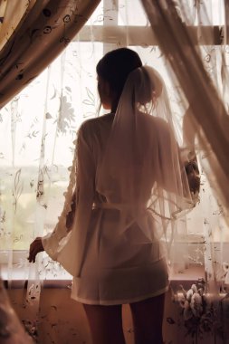 Kırsal evdeki pencerede bornozlu sevimli genç bir gelin. Gelinin düğün gününün sabahı. Mutlu dişi damadı ile tanışmak için bekliyor. Mutlu ve lüks evlilik kavramı. Alan için kopyala