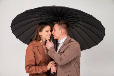 Siyah şemsiyeli genç ve şık bir çift aşk duyguları gösteriyor. Güz giysileri içinde açık gri arka plana bakarken şemsiye altında duran sevimli duygusal insanların portresi. Alan için kopyala