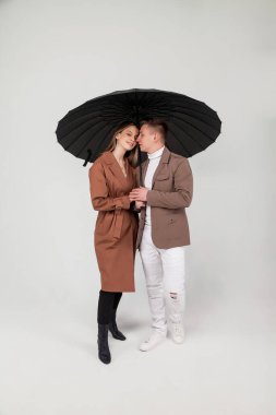 Siyah şemsiyeli genç ve şık bir çift aşk duyguları gösteriyor. Güz giysileri içinde açık gri arka plana bakarken şemsiye altında duran sevimli duygusal insanların portresi. Alan için kopyala