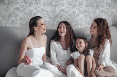 Three adult women and small girl in stylish casual white clothes spend time on bed, chat sweetly, tell stories and laugh. Friends met and spend their leisure time in hotel room. Concept of friendship