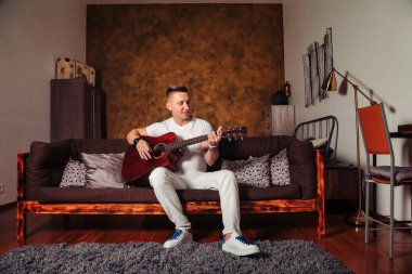Beyaz elbiseli, gitarlı harika bir genç adam oturma odasının içinde. Kanepede gitar çalan sevimli bir erkek. Evde öğrenme ya da müzikal çalma kavramı ve müzik eğitimi. Alan için kopyala