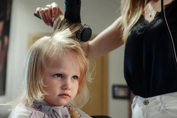 Barber woman make fashionable pretty hairstyle for cute little blond girl child in modern barbershop, hair salon. Hairdresser makes hairdo for young baby in barber shop. Concept hairstyle and beauty