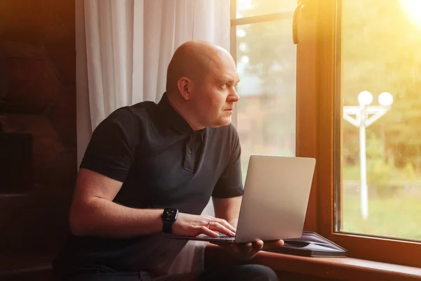 Portrait of young businessman with laptop at window of living room in country house. Workaholic man in home casual clothes working on vacation. Creative inspiration and start-up business. Copy space