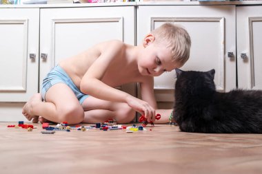 5-7 yaşlarında bir çocuk evde inşaat setinde, siyah bir kedinin yanında oynuyor.