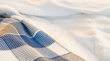 Mavi ve gri renkli kareli battaniye yumuşak ışıklı bir yatak odasındaki beyaz yatağa dayanır.