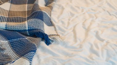 Mavi ve bej renkli kareli battaniye görünür desenli beyaz bir yatak örtüsünün üzerine örtülmüş.