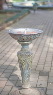 Yeşilliklerle çevrili halka açık bir parkta, taştan bir patikada duran süslü metal çeşme.