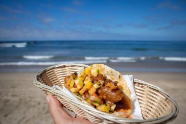 A hand holding a fish taco in basket in front of an ocean landscape in Mexico clipart