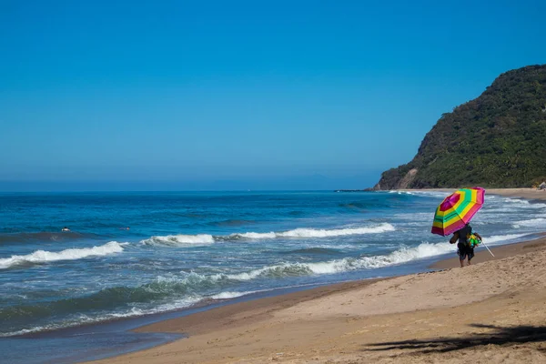 Lo de Marcos, Nayarit, Meksika - 23 Ocak 2021: Bir adam Meksika 'nın Riviera Nayarit sahilinde elinde gökkuşağı şemsiyesi ile kumlu sahilde yürüyor