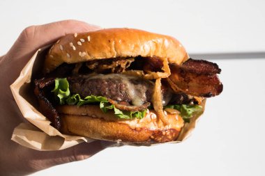 Bir el taze pişmiş, sulu bir batı barbekü burgeri, peynirli, kızarmış, soğanlı, marullu, pastırmalı, barbekü soslu beyaz arka planda susamlı bir çörek..