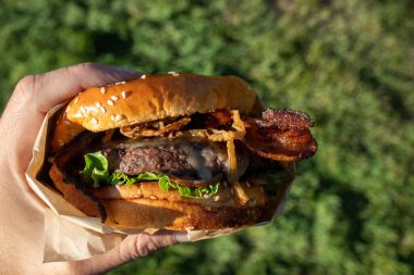 Bir el taze pişmiş, sulu bir batı barbekü burgeri, peynirli, kızarmış, soğanlı, marullu, pastırmalı, barbekü soslu yeşil çimenli bir arka planda susamlı çöreğin üstünde..