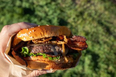 Bir el taze pişmiş, sulu bir batı barbekü burgeri, peynirli, kızarmış, soğanlı, marullu, pastırmalı, barbekü soslu yeşil çimenli bir arka planda susamlı çöreğin üstünde..