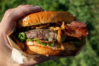 Bir el taze pişmiş, sulu bir batı barbekü burgeri, peynirli, kızarmış, soğanlı, marullu, pastırmalı, barbekü soslu yeşil çimenli bir arka planda susamlı çöreğin üstünde..